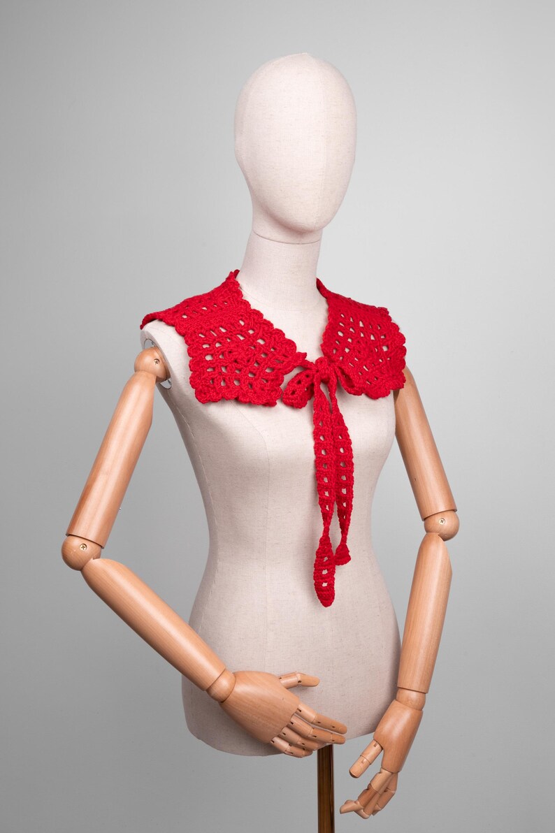 May include: A bright red, crocheted collar with a scalloped edge and long, tie-up closures. The collar is displayed on a mannequin with wooden arms and hands, against a neutral gray background. The collar is a handmade accessory.