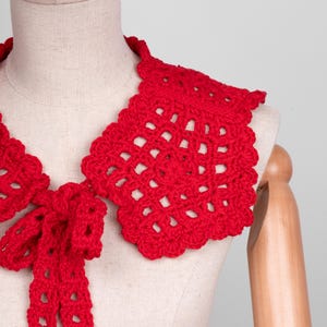 May include: A bright red, handmade crochet collar with a scalloped edge and a bow tie. The collar is displayed on a mannequin, showcasing its openwork design and vibrant color. This accessory adds a touch of vintage charm.