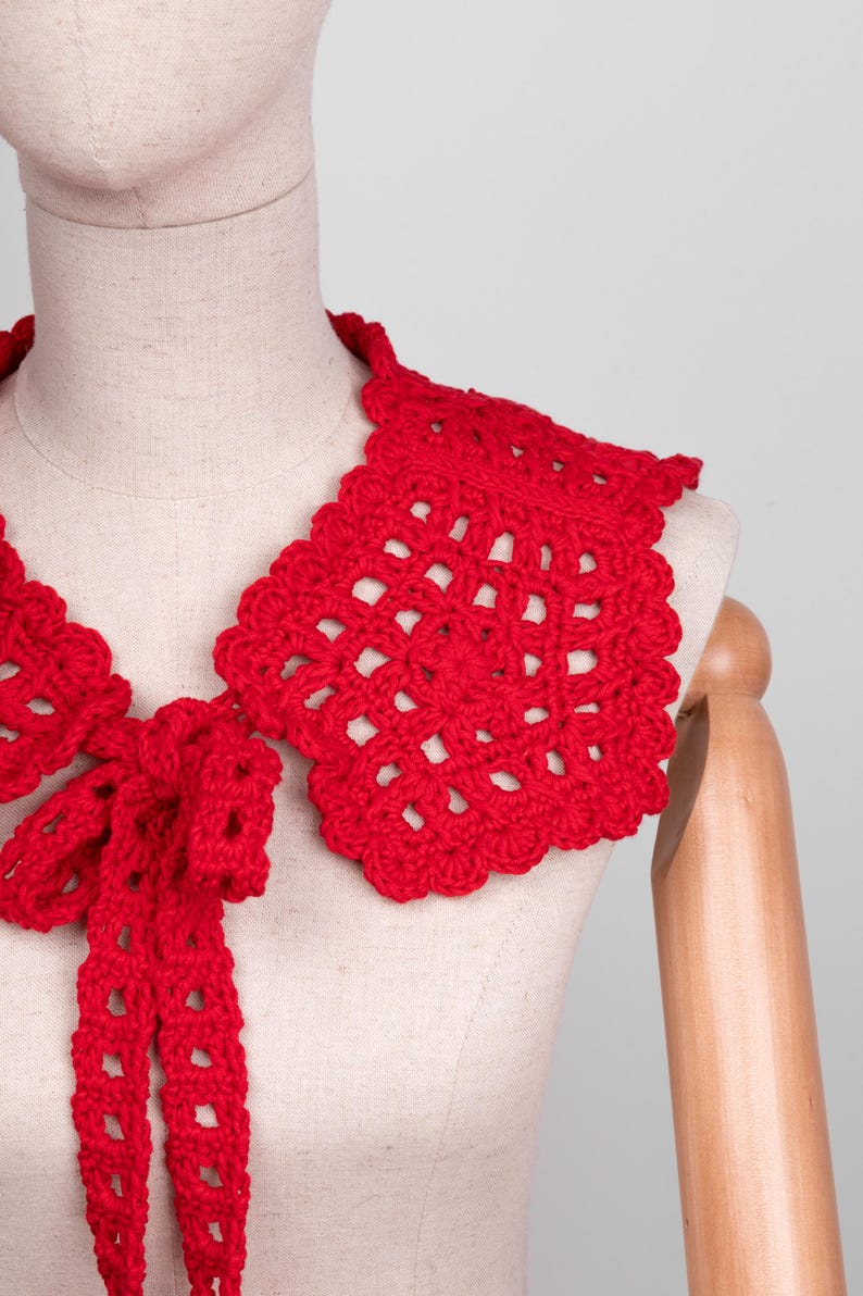 May include: A bright red, handmade crochet collar with a scalloped edge and a bow tie. The collar is displayed on a mannequin, showcasing its openwork design and vibrant color. This accessory adds a touch of vintage charm.