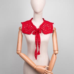 May include: A bright red, crocheted collar with a scalloped edge and a tie closure. The collar is displayed on a mannequin, showcasing its openwork design and the long, dangling ties of the bow.