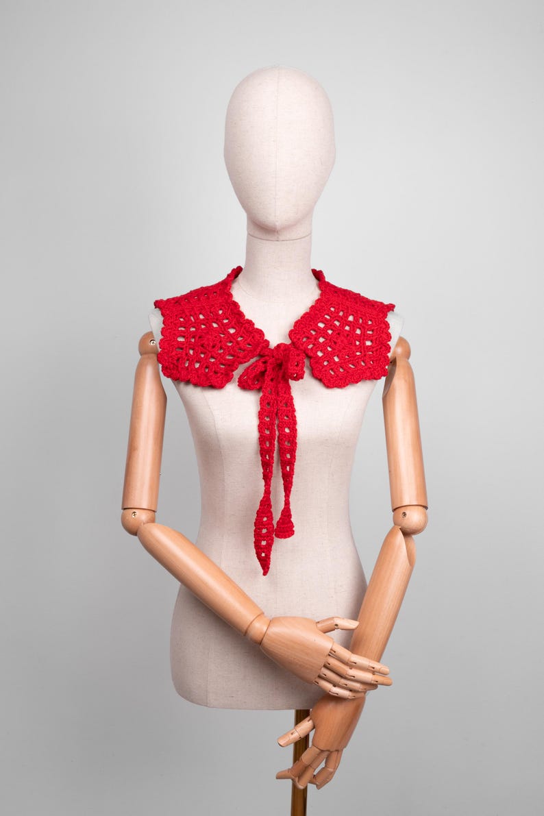 May include: A bright red, crocheted collar with a scalloped edge and a tie closure. The collar is displayed on a mannequin, showcasing its openwork design and the long, dangling ties of the bow.