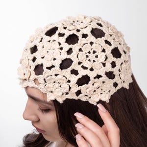 Puede incluir: Una diadema de ganchillo de color crema con un diseño floral. El patrón calado permite vislumbrar el cabello que hay debajo. La diadema está hecha de un hilo suave y texturizado y se lleva en la cabeza.