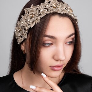 K&ouml;nnte beinhalten: Ein handgefertigtes, beigefarbenes H&auml;kel-Stirnband mit Blumen- und Sternenmustern. Das Stirnband ist mit kleinen, funkelnden Verzierungen versehen. Das Accessoire wird von einer Person mit langen, braunen Haaren und einem schwarzen Oberteil getragen.