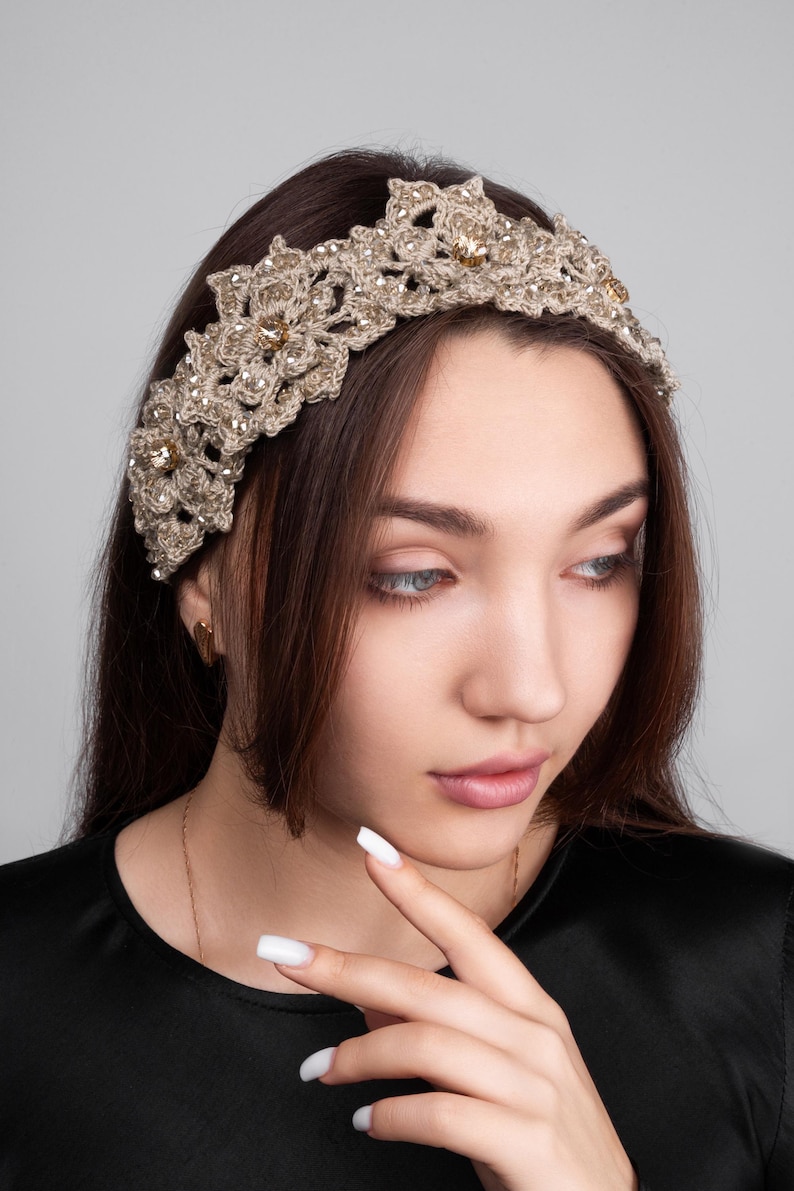 K&ouml;nnte beinhalten: Ein handgefertigtes, beigefarbenes H&auml;kel-Stirnband mit Blumen- und Sternenmustern. Das Stirnband ist mit kleinen, funkelnden Verzierungen versehen. Das Accessoire wird von einer Person mit langen, braunen Haaren und einem schwarzen Oberteil getragen.