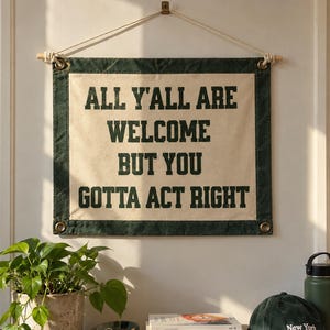 May include: A rectangular canvas wall hanging with a dark green border and cream center. The text in dark green block letters reads: ALL Y'ALL ARE WELCOME BUT YOU GOTTA ACT RIGHT. It is suspended from a wooden dowel with a white cord for hanging.