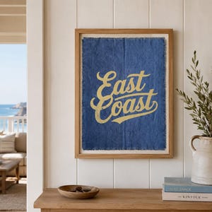 Op de afbeelding: Een ingelijst kunstwerk aan de muur met een gestructureerde blauwe stoffen achtergrond waarop de woorden East Coast in een gouden scriptlettertype zijn gedrukt. Het kunstwerk zit in een licht houten frame en heeft gerafelde randen.