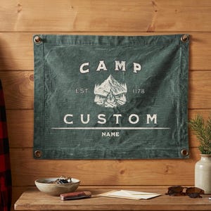 Peut inclure: Une bannière murale rectangulaire en toile vert foncé avec un graphique blanc représentant une montagne et un feu de camp. Le texte indique CAMP, EST 1178, CUSTOM et NAME. La bannière possède des œillets métalliques aux coins.