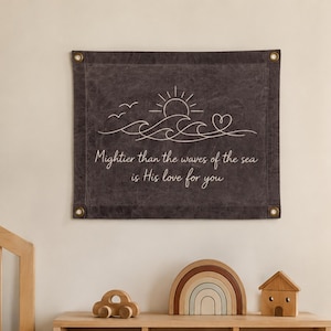 May include: A dark gray rectangular wall hanging featuring a white line art illustration of a sun, waves, birds, and a heart. The text reads: Mightier than the waves of the sea is His love for you. The fabric has four metal grommets at the corners.