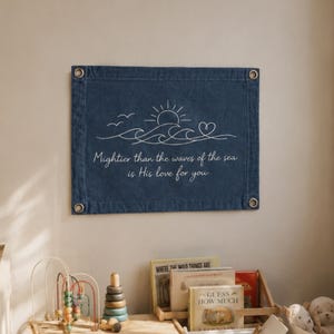 Puede incluir: Un tapiz rectangular de mezclilla azul oscuro con arte lineal blanco de un sol, olas, pájaros y un corazón. El texto dice: "Mightier than the waves of the sea is His love for you." La tela tiene cuatro ojales metálicos en las esquinas.