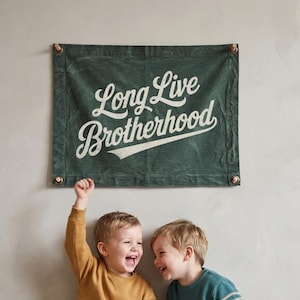 May include: A green banner with the text "Long Live Brotherhood" hangs above two young boys. One boy wears a mustard-colored sweater and jeans, while the other wears a striped teal sweater and brown pants. Both are laughing.
