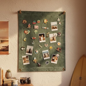 Könnte beinhalten: Eine grüne Leinwand-Memo-Tafel mit Fotos und dekorativen Pins. Die Tafel hängt an einer weißen Wand, mit einem Holztisch, Büchern, einer Tasse und einer Vintage-Kamera im Vordergrund. Ein Surfbrett lehnt an der Wand.