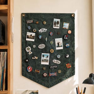 Peut inclure: Une bannière murale en toile vert foncé avec un bord inférieur pointu et deux œillets métalliques en haut. Le tissu est recouvert de diverses épinglettes en émail, de petits badges circulaires et de plusieurs photos instantanées carrées.