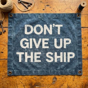 May include: A blue banner with the text "DON'T GIVE UP THE SHIP" in white. The banner has grommets in each corner and is displayed on a wooden surface. Decorative items like keys and twine are also visible.