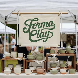 Könnte beinhalten: Ausstellung handgefertigter Keramikbecher, Schalen und einer Vase in Weiß-, Grün- und Brauntönen. Über der Ausstellung hängt ein Banner mit der Aufschrift "Forma Clay". Die Artikel sind auf Holzregalen angeordnet.