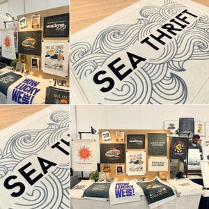 May include: A collection of rectangular fabric wall banners featuring various graphic designs and text, including the phrase SEA THRIFT in black lettering over a blue wave pattern, displayed on a wooden board and a white table surface.