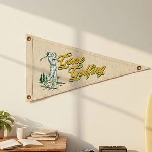May include: A beige pennant flag with the words "Gone Golfing" in yellow script. The flag features a green and white illustration of a golfer swinging a club. The pennant has three metal grommets.