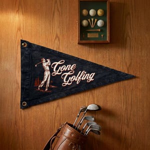 May include: A navy blue pennant with the words "Gone Golfing" in white script, featuring a golfer illustration. A brown leather golf bag with clubs is at the bottom. A framed display of golf balls and tees is at the top.