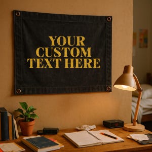 May include: A rectangular black fabric wall banner with metal grommets in each corner, featuring the yellow distressed text YOUR CUSTOM TEXT HERE printed in the center. The banner is displayed on a wall above a wooden desk with various items.
