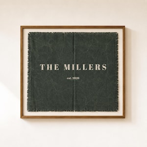 Könnte beinhalten: Ein quadratischer Wandbehang aus dunkelgrünem Stoff mit ausgefransten Kanten in einem hellbraunen Holzrahmen. Der Stoff zeigt den weißen Schriftzug THE MILLERS zentriert über dem kleineren Text est. 2020 auf einer strukturierten, knittrigen Oberfläche.