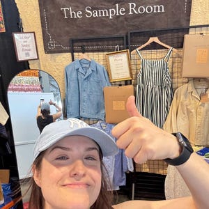 May include: A display featuring a dark banner with the text The Sample Room, a light blue collared jacket, a black and white striped dress, and a tan shirt. A small sign reads Ask About our Sewing Retreats and another reads Pattern Drafting.