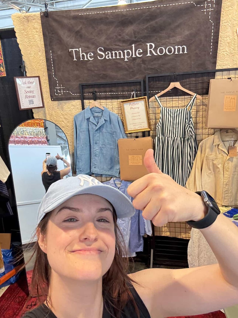 May include: A display featuring a dark banner with the text The Sample Room, a light blue collared jacket, a black and white striped dress, and a tan shirt. A small sign reads Ask About our Sewing Retreats and another reads Pattern Drafting.