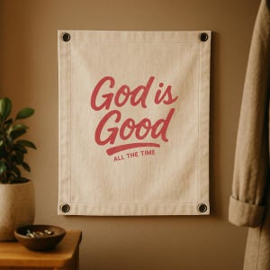 Op de afbeelding: Een beige canvas banner met de woorden "God is Good" in rood script, met "ALL THE TIME" eronder. De banner hangt aan een bruine muur met metalen ogen in elke hoek.