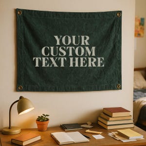 Könnte beinhalten: Dunkelgrüner Wandbehang aus Canvas mit dem Text "YOUR CUSTOM TEXT HERE" in Weiß. Das Banner hat Metallösen in jeder Ecke. Das Banner hängt an einer Wand über einem Schreibtisch mit einer Lampe, Büchern und einer kleinen Topfpflanze.