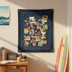 Op de afbeelding: Een donkerblauw stoffen memobord met diverse kleurrijke spelden en stickers, waaronder afbeeldingen van golven, regenbogen en palmbomen. Een houten bureau met een mok, camera en luidspreker staat op de voorgrond. Een surfplank leunt tegen de muur.