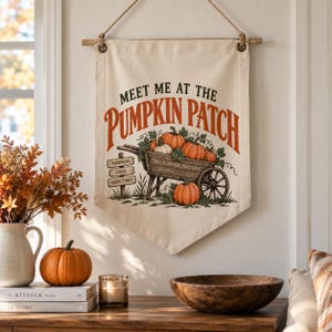 Peut inclure: Une tenture murale en toile couleur crème ornée d'un graphique représentant une brouette en bois remplie de citrouilles orange. Le texte indique "MEET ME AT THE PUMPKIN PATCH" en noir et orange, avec un panneau indiquant "PUMPKINS", "LOCAL", "GOOD TIMES".