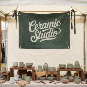 Op de afbeelding: Een display van keramische mokken, kommen en vazen op houten verhogingen, op een wit tafelkleed. Een groene banner met de tekst "Ceramic Studio" hangt erboven. Het aardewerk is in tinten groen, grijs en wit.