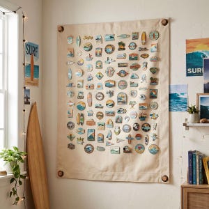 Pode incluir: Um grande tecido bege pendurado na parede com inúmeros patches com tema de surf. Os patches apresentam vários designs, incluindo pranchas de surf, ondas e imagens costeiras. A palavra "SURF" é visível em um pôster e um patch.