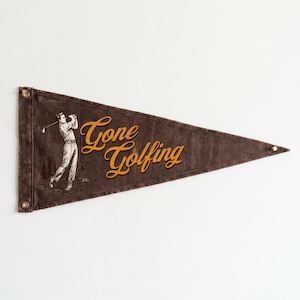 May include: A brown pennant with the words "Gone Golfing" in gold script. A vintage illustration of a golfer swinging a club is on the left side. The pennant has metal grommets for hanging.