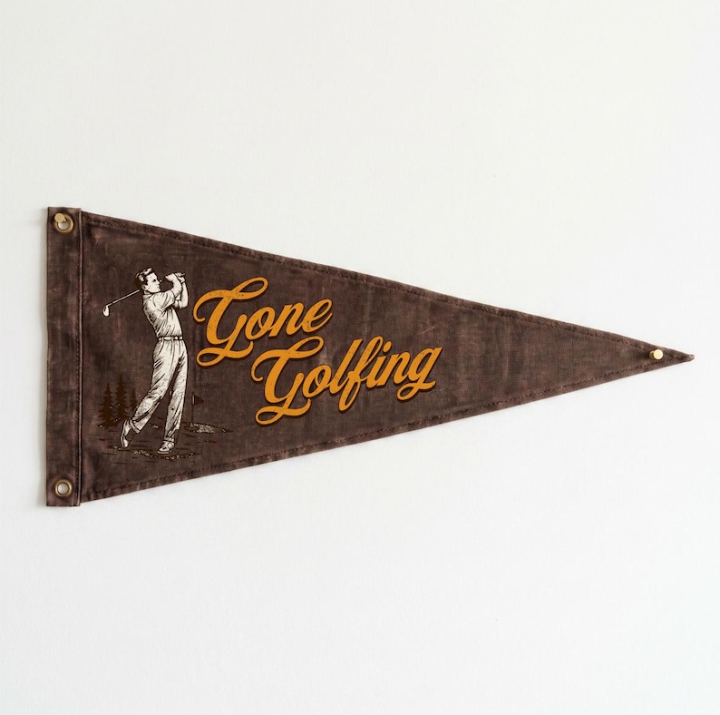 May include: A brown pennant with the words "Gone Golfing" in gold script. A vintage illustration of a golfer swinging a club is on the left side. The pennant has metal grommets for hanging.