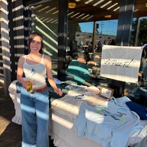 May include: A cream-colored tank top with a small embroidered tree design, blue denim jeans with a wide-leg cut, and a table display featuring light blue and cream sweatshirts with whale and tree graphics and a banner reading Le Petit Fil.