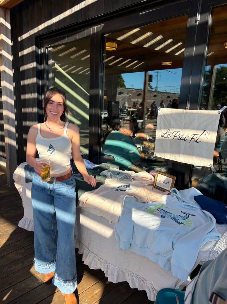 May include: A cream-colored tank top with a small embroidered tree design, blue denim jeans with a wide-leg cut, and a table display featuring light blue and cream sweatshirts with whale and tree graphics and a banner reading Le Petit Fil.