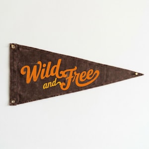 Peut inclure: Un fanion triangulaire marron foncé avec le texte orange Wild and Free. Le fanion possède deux œillets métalliques sur le côté gauche pour la suspension et une pointe sur le côté droit, présentant une texture usée sur toute la surface.