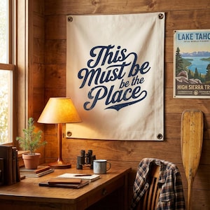 Può includere: Un arazzo da parete in tela con la scritta "This Must be the Place" in blu navy è esposto su una parete di legno. Sotto c'è una scrivania con una lampada, libri e un piccolo albero in vaso. È visibile anche un poster vintage del lago Tahoe.