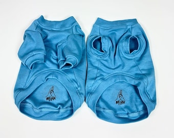 May include: Two light blue dog shirts are displayed on a white surface. The shirts have long sleeves and openings for the legs. Each shirt has a black graphic near the bottom. The shirts are made of a soft material.