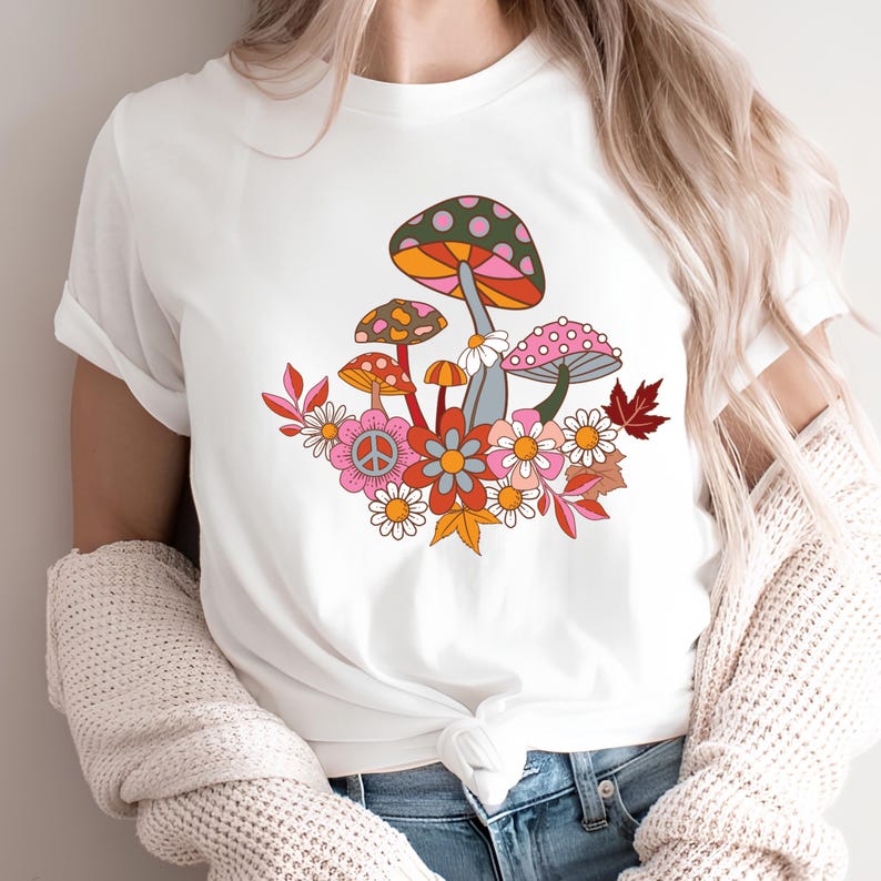 May include: A white short-sleeved t-shirt featuring a colorful graphic print of various mushrooms, daisies, and autumn leaves. The design includes a peace sign symbol within a flower and a palette of orange, pink, green, and red colors.