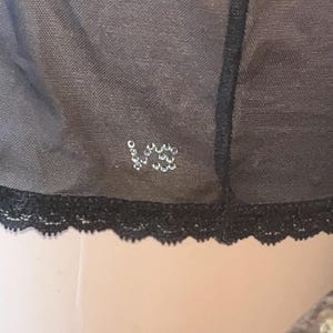 May include: Close-up of a black lingerie item with a delicate lace trim. The garment features a small, sparkling "VS" logo made of rhinestones. The fabric appears to be a sheer, dark material.