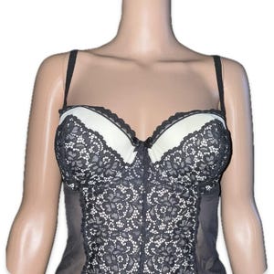 May include: A black and white lace bustier with adjustable straps. The bustier has white padded cups with black lace trim and a black lace overlay on the bodice. The garment is displayed on a mannequin.
