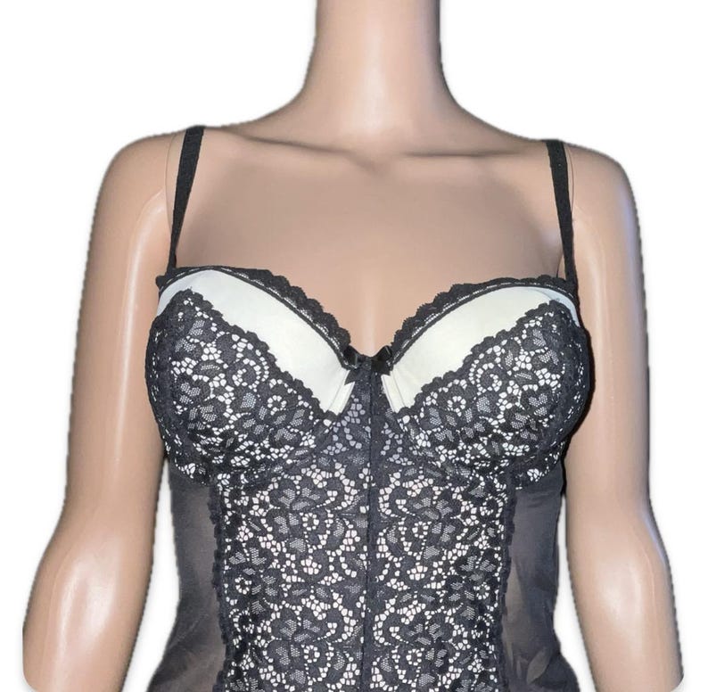 May include: A black and white lace bustier with adjustable straps. The bustier has white padded cups with black lace trim and a black lace overlay on the bodice. The garment is displayed on a mannequin.