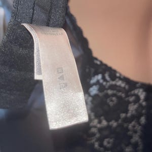 May include: Close-up of a black bra with a lace design. A white tag is attached, displaying care symbols. The bra's fabric appears soft, and the overall aesthetic is delicate and feminine. The tag is rectangular and has a slightly reflective surface.