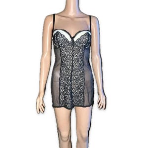 May include: Black lingerie chemise with a white bust and black lace detailing. The chemise has thin straps and a sheer skirt. The lace design is centered on the front of the chemise.