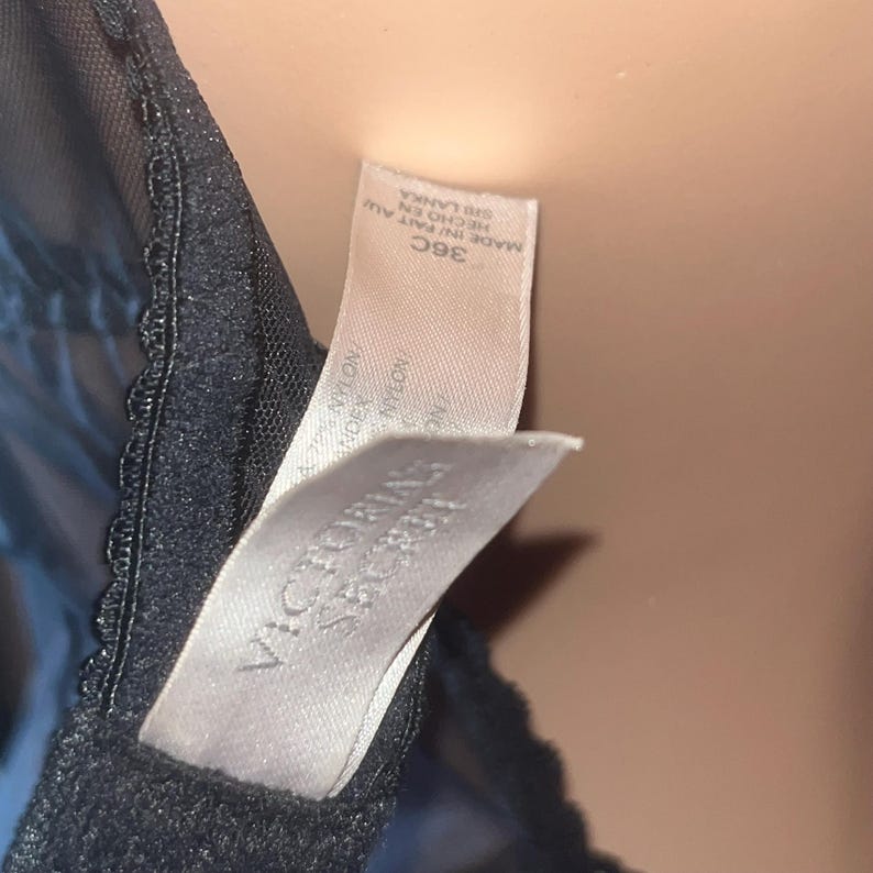 May include: Close-up of a black bra with a lace trim. The bra has a tag that reads "VICTORIA'S SECRET" and "36C". The tag also indicates the bra was made in Sri Lanka.