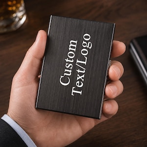 May include: A rectangular black metal card holder with a brushed finish. The front surface features the white engraved text Custom Text/Logo oriented vertically. The object has a slim profile with a metallic side hinge mechanism visible on the edge.