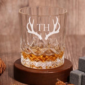 May include: A clear glass whisky tumbler with a cut glass design. The glass has an etched design of antlers and the initials "TH". The glass is sitting on a wooden coaster.