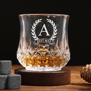 Könnte beinhalten: Ein klares, graviertes Whiskyglas mit dem Text "BEST MAN" und dem Datum 9.12.2024, gefüllt mit bernsteinfarbener Flüssigkeit. Das Glas steht auf einem Holzuntersetzer, mit drei grauen Eiswürfeln und einem Tannenzapfen in der Nähe.
