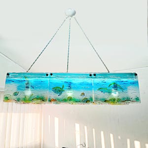 May include: A rectangular, hanging light fixture with a marine theme. The fixture features a blue and green underwater scene with dolphins, sea turtles, and fish. The light is suspended by three silver wires from a white ceiling fixture.