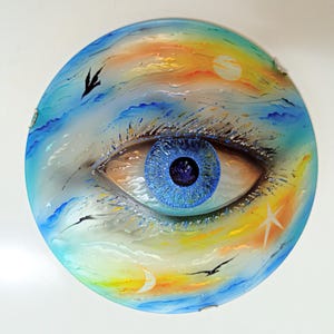 May include: A round glass wall art piece with a blue eye painted on it. The eye is surrounded by a blue, white, and yellow sky with birds flying. The background also includes a crescent moon and a star.